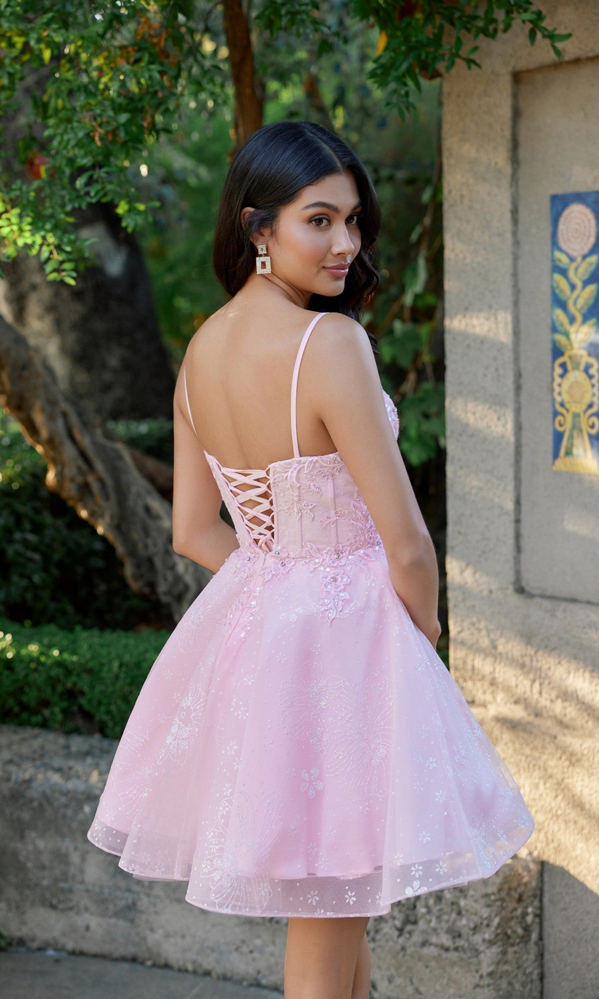 Wearing the Juliet JT985L, a light pink glitter tulle homecoming dress with a lace-up back and corset bodice, a woman stands outdoors near a stone wall and greenery, glancing over her shoulder.